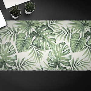 May include: A white desk mat featuring a tropical leaf design. The leaves are green and have a realistic texture. The mat is ideal for adding a touch of nature to your workspace.
