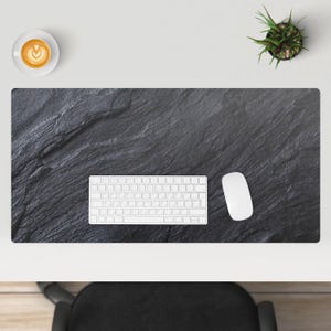 Puede incluir: Una alfombrilla de escritorio negra con una superficie texturizada, un teclado blanco y un ratón blanco. Una taza de café con arte latte y una pequeña planta en maceta también son visibles en la superficie blanca.