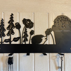 May include: Black metal key holder shaped like a hedgehog in a pot with flowers and leaves. The key holder has five hooks for hanging keys.