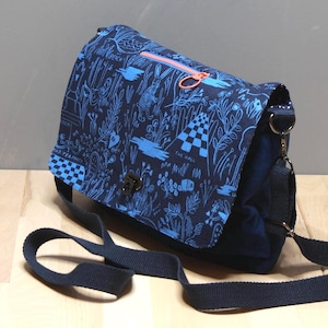 Könnte beinhalten: Dunkelblaue Messenger-Tasche mit einem skurrilen blau-weißen, illustrierten Muster. Die Tasche hat einen leuchtend orangefarbenen Reißverschluss und einen schwarzen, verstellbaren Riemen. Die Klappe hat ein Schachbrettmuster und die Worte "THE HALL" sind sichtbar.