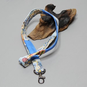 Könnte beinhalten: Ein Stoff-Schlüsselband mit blauem und Paisley-Muster, mit einem silberfarbenen Metallverschluss. Das Schlüsselband wird auf einer grauen Oberfläche mit einem Stück Treibholz im Hintergrund präsentiert.