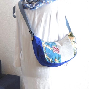 Könnte beinhalten: Eine Umhängetasche mit blauem Riemen und floralem Muster. Die Tasche hat eine geschwungene Form, mit einem blauen, samtartigen Material am Boden und einem Blumendesign in der Mitte. Ein passender floraler Schal ist über der Schulter drapiert.