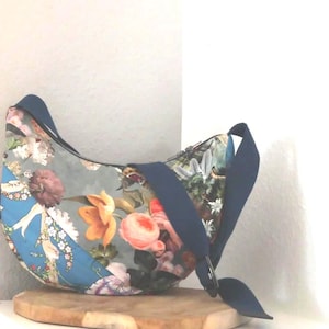 Könnte beinhalten: Eine halbmondförmige Umhängetasche mit einem lebendigen Blumenmuster in Blau-, Orange-, Rosa- und Grüntönen. Die Tasche verfügt über einen einfarbigen, dunkelblauen, verstellbaren Riemen mit einem Metallring und einen Reißverschluss oben.