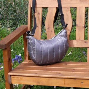 Könnte beinhalten: Eine silberne, gesteppte Umhängetasche mit einem verstellbaren schwarzen Riemen liegt auf einer Holzbank. Die Tasche hat eine gebogene Form und einen Reißverschluss. Die Bank ist aus Holz und befindet sich im Freien in einem Garten.