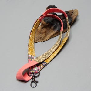 Könnte beinhalten: Ein gemustertes Schlüsselband mit einem korallenfarbenen Nackenband und einem silberfarbenen Metallverschluss. Das Schlüsselband hat ein Blumen- und geometrisches Design in Gelb-, Gold- und Grautönen.