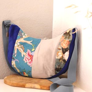 Könnte beinhalten: Eine halbmondförmige Umhängetasche mit einem Patchwork-Design aus blauem Samt, einem türkisfarbenen Paneel mit Blumen- und Astmuster, einem beigen Mittelteil und einem Blumenmuster-Paneel mit orangefarbenen und weißen Blüten sowie einem grauen Gurt.