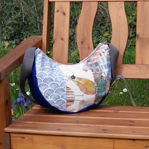 Könnte beinhalten: Eine halbmondförmige Umhängetasche mit grauem Riemen liegt auf einer Holzbank. Die Tasche hat ein blau-weißes Wellenmuster, eine Vogelillustration und ein Stadtbilddesign. Der Besatz der Tasche ist tiefblau.