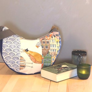 Könnte beinhalten: Eine halbmondförmige Umhängetasche mit blauem Riemen und farbenfrohem Design. Die Tasche zeigt eine Vogelillustration und ein Stadtbildmuster. Ein Buch, eine Tasse und ein grüner Kerzenhalter befinden sich neben der Tasche.