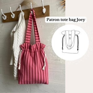May include: A pink and maroon striped tote bag with matching straps. The bag has a drawstring closure and white tassels. The text "Patron tote bag Joey" is visible in the image. A white sweater hangs on a hook in the background.