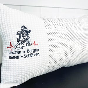 Könnte beinhalten: Weißes rechteckiges Kissen mit Waffelstruktur auf einer Seite und einem gepunkteten Muster auf der anderen. Mit gestickter Feuerwehrgrafik und deutschem Text auf der Waffelseite.