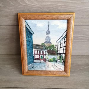 May include: A framed oil painting depicting a narrow street in a European town. The street is lined with buildings, and a church steeple rises in the distance. The painting is signed and dated "S. Schneider 2002".