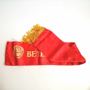 May include: A red satin sash with gold fringe and gold lettering that reads "VETERAN". The sash features a gold emblem with a hammer and sickle design.