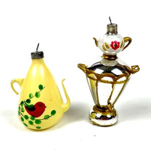 Puede incluir: Dos adornos navideños vintage. Uno es una tetera amarilla con un pájaro rojo y hojas verdes. El otro es un samovar plateado y dorado con una parte superior blanca y un diseño de flor roja. Ambos tienen tapas metálicas.
