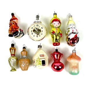 Op de afbeelding: Een verzameling vintage kerstversieringen in verschillende vormen en kleuren. Bevat een klok, een sneeuwpop, een huis en andere decoratieve figuren. Elke versiering heeft een metalen dop om op te hangen.