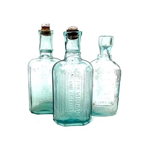 May include: Three antique, light blue glass bottles with cork stoppers. The bottles have a rectangular shape with vertical ridges and embossed text. The bottles are displayed against a white background.