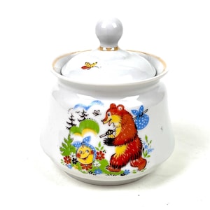 May include: White ceramic sugar bowl with a lid. The bowl features a colorful illustration of a bear carrying a bag, a smiling sun, and floral accents. The lid has a small knob and a gold rim.