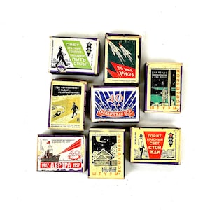 May include: A collection of vintage matchboxes with various designs and text in multiple languages. The boxes feature illustrations of rockets, buildings, and figures, with a color palette including red, green, and blue.