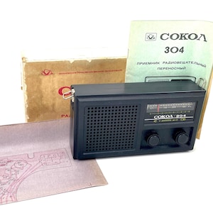 Könnte beinhalten: Ein schwarzes Vintage-Radio Sokol 304 mit Lautsprechergitter, zwei Knöpfen und einem Abstimmungsrad. Das Radio wird von seiner Originalverpackung, einer Bedienungsanleitung und einem Schaltplan begleitet. Der Text "SOKOL 304" ist auf dem Radio und der Anleitung sichtbar.