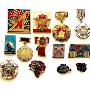 May include: A selection of vintage enamel pins and badges, featuring diverse designs with red, gold, and blue hues. The pins showcase stars, ships, and architectural elements, with text including the year "1917".