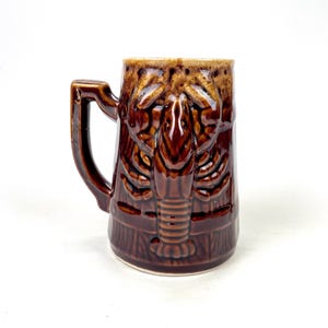 May include: A brown ceramic beer mug with a detailed lobster design. The mug has a handle and a textured surface, with a darker brown glaze on the lobster and a lighter brown glaze on the rim.