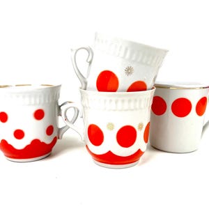 Puede incluir: Cuatro tazas de cerámica blanca con lunares rojos y ribete dorado. Las tazas tienen asas y un diseño festoneado decorativo. Una taza tiene un diseño de estrella. Las tazas están apiladas sobre un fondo blanco.