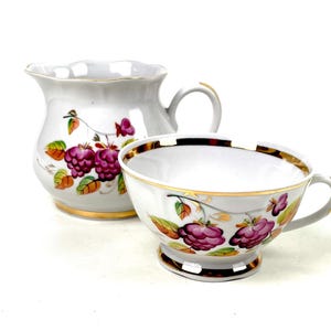May include: A white porcelain creamer and teacup set. The set features a gold rim and is decorated with a floral design of purple raspberries and green leaves. The creamer has a handle and a scalloped edge.