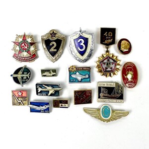 May include: A collection of vintage Soviet-era pins and badges. The pins feature various designs, including stars, airplanes, and military emblems. Colors include red, gold, blue, and white. Some pins have text in Cyrillic script.