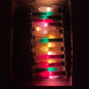 May include: A string of electric candle lights arranged in a cardboard box. The set features multiple plastic candle-shaped bulbs in various colors including red, green, yellow, and white, connected by black wiring. Each bulb has a flame-like tip.