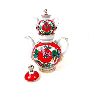 May include: A white porcelain teapot and a smaller teapot stacked on top, both with a vibrant red floral design. The teapots have gold accents and a matching lid. The design features red flowers, green leaves, and pink accents.