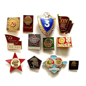 Może przedstawiać: Kolekcja zabytkowych przypinek i odznak z epoki sowieckiej. Przypinki mają różne kształty, kolory i wzory, w tym gwiazdy, flagi i portrety. Niektóre przypinki mają tekst w cyrylicy.