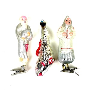 Puede incluir: Tres adornos navideños vintage de cristal. Uno es una figura con una túnica plateada sosteniendo un globo, otro es un pez con detalles rojos, y el tercero es una figura con una túnica blanca con ribetes rojos.