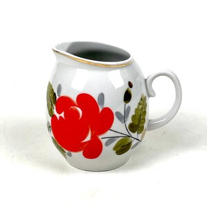 Pode incluir: Cremeira de cerâmica branca com um design floral vermelho. A cremeira tem um bico curvo, uma alça e uma borda dourada. O design floral inclui flores vermelhas, folhas verdes e caules cinzentos. Uma peça decorativa para servir creme ou leite.