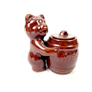 May include: A brown ceramic bear figurine holding a matching barrel. The barrel has a lid and the word "HONEY" is embossed on the side. The bear is glazed and has a glossy finish. The figurine is a decorative item.