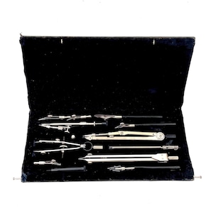 Pode incluir: Um conjunto de ferramentas de desenho, incluindo compassos e divisores, dispostos em um estojo forrado de veludo preto. As ferramentas são feitas de metal prateado e plástico preto. O estojo aberto revela os instrumentos, sugerindo precisão e design.