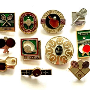 May include: A selection of vintage tennis pins in diverse shapes and designs. The pins depict tennis rackets, balls, and nets, with text in a foreign language. The colour scheme includes green, red, gold, and white, with some pins having a circular or rectangular form.