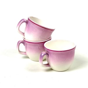 May include: Four stacked teacups with a white interior and a pink ombre exterior. Each cup has a handle and a thin, dark pink rim. The cups are arranged on a white background.