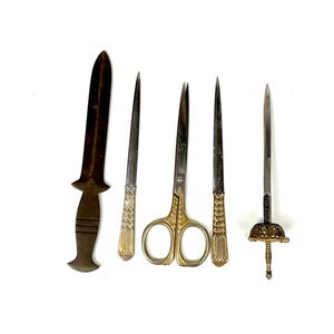 Może przedstawiać: Asortyment antycznych otwieraczy do listów i nożyczek. Kolekcja obejmuje brązowy otwieracz do listów, dwa srebrne otwieracze do listów, złote nożyczki i ozdobny otwieracz do listów w kształcie miecza z napisem "TOLEDO" na ostrzu.