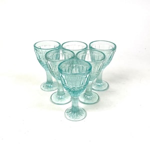 Puede incluir: Seis copas de vino vintage de color azul claro. Cada copa presenta un diseño geométrico texturizado en el cuenco y el tallo. Las copas son de vidrio transparente. Dispuestas en un patrón circular.