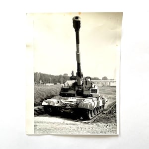 Puede incluir: Fotografía en blanco y negro de un tanque militar. El tanque tiene un cañón largo apuntando hacia arriba. El tanque está camuflado con una mezcla de colores oscuros y claros. El tanque está en un camino de tierra.