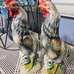 May include: Two ceramic rooster figurines standing on textured bases. Each rooster features a grey and white feathered body, a bright red comb and wattle, and yellow legs. The birds are depicted in an upright, alert posture with detailed plumage textures and painted highlights throughout their bodies.