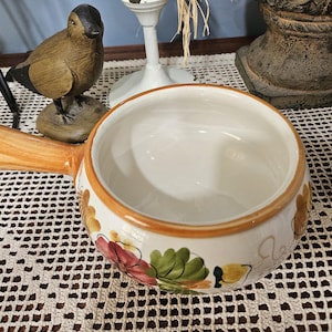 Può includere: Una zuppiera in ceramica bianca con manico, con un motivo floreale rosso, verde e giallo. Il bordo e il manico sono dipinti di arancione. Una statuetta decorativa di uccello e altri oggetti sono sullo sfondo.