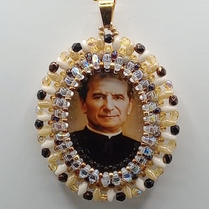 Puede incluir: Colgante ovalado con un retrato de un hombre con traje negro, rodeado por un marco de cuentas. El marco incluye cuentas blancas, doradas y negras, con una cadena dorada.