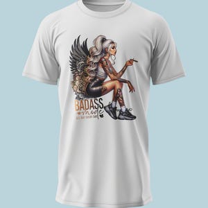 Könnte beinhalten: Ein weißes Kurzarm-T-Shirt mit einer Grafik einer tätowierten Figur mit langem blonden Haar und dunklen Flügeln, die sitzt und einen kleinen Gegenstand hält. Der Text BADASS mode ALL DAY EVERY DAY steht in schwarzer und goldener Schrift vorne.