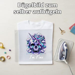 Könnte beinhalten: Weißes T-Shirt mit einem farbenfrohen Grafikdesign eines Schädels mit einem Einhornhorn, umgeben von Wolken und Tränen, mit dem Text "I'm Fine". Ein Bügeleisen und eine Schere liegen neben dem Shirt.
