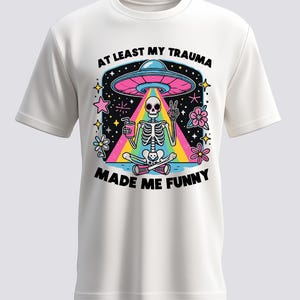 Könnte beinhalten: Weißes T-Shirt mit einer bunten Grafik eines Skeletts, das unter einem UFO meditiert. Das Skelett hält ein Getränk und zeigt das Friedenszeichen. Der Text lautet "AT LEAST MY TRAUMA MADE ME FUNNY."