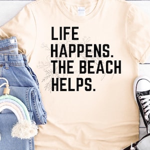 May include: A beige t-shirt with the text "Life Happens. The Beach Helps." printed in black. The shirt is laid out on a white wooden surface with a pair of blue jeans and a rainbow-colored yarn rainbow hanging on the jeans. A pair of black canvas shoes are on the right side of the image.