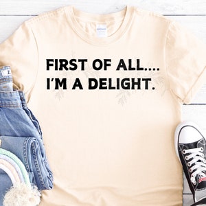 Könnte beinhalten: Ein beiges T-Shirt mit dem Text "FIRST OF ALL... I'M A DELIGHT." in Schwarz gedruckt.