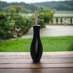 Puede incluir: Un jarrón difusor negro de estilo moderno con un diseño abierto único. El jarrón contiene varias varillas de madera y está colocado sobre una superficie de madera con un fondo verde borroso.