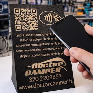 Può includere: Un espositore nero angolato con un codice QR e le parole "lascia una recensione" in più lingue. L'espositore presenta il logo Doctor Camper, il numero di telefono e l'indirizzo del sito web. Uno smartphone è tenuto vicino all'espositore.