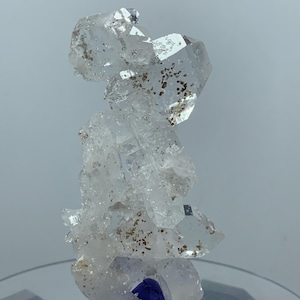 May include: A cluster of clear, translucent quartz crystals featuring sharp, angular points and facets. The mineral specimen contains small, scattered brown inclusions throughout its structure, resting on a small, dark blue base.